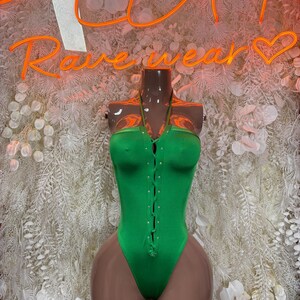 May include: A bright green bodysuit with a halter neckline and a series of small, decorative buttons down the front. The bodysuit is displayed on a mannequin. The neon sign above reads "Rave Wear".