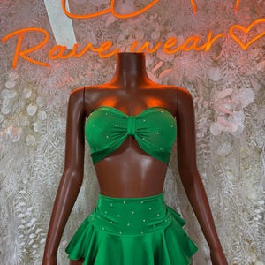 May include: A green two-piece outfit with rhinestone embellishments. The top is a strapless bandeau style with a bow detail. The skirt is high-waisted with a ruffled hem. The outfit is displayed on a headless mannequin.