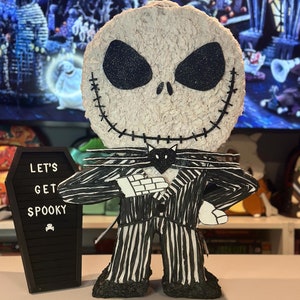 Puede incluir: Una pi&ntilde;ata de Jack Skellington a rayas blancas y negras con una pajarita negra y una cara de gato negro. La pi&ntilde;ata est&aacute; de pie sobre una mesa frente a un ata&uacute;d negro con las palabras "Let's Get Spooky" escritas en &eacute;l.
