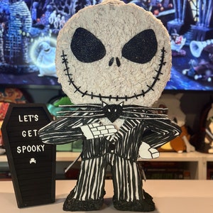 Puede incluir: Una figura de Jack Skellington con rayas blancas y negras, con una pajarita negra y un gato negro en el pecho. La figura est&aacute; de pie frente a una pizarra negra con el texto "Let's Get Spooky" escrito en ella.
