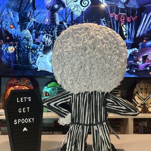Puede incluir: Una pi&ntilde;ata de Jack Skellington a rayas blancas y negras con una cabeza blanca y esponjosa. La pi&ntilde;ata est&aacute; de pie sobre una mesa frente a una pizarra negra con el texto "Let's Get Spooky" escrito en ella.