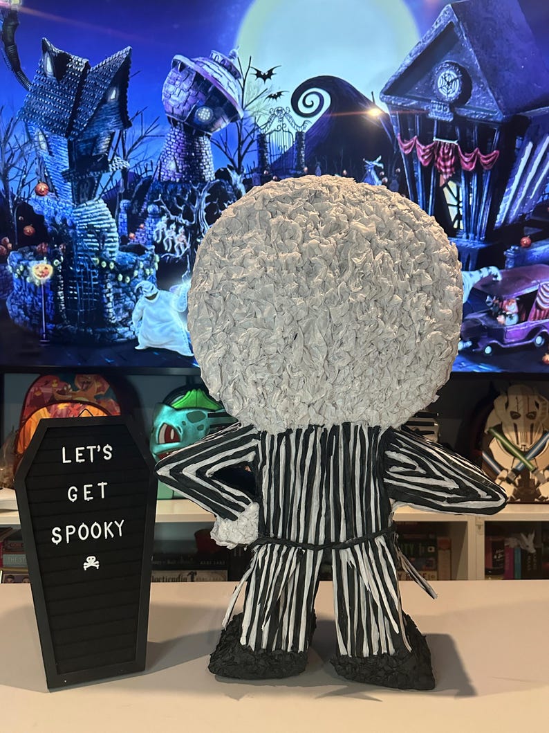 Puede incluir: Una pi&ntilde;ata de Jack Skellington a rayas blancas y negras con una cabeza blanca y esponjosa. La pi&ntilde;ata est&aacute; de pie sobre una mesa frente a una pizarra negra con el texto "Let's Get Spooky" escrito en ella.