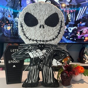 Puede incluir: Una pi&ntilde;ata de Jack Skellington en blanco y negro con un traje a rayas y una pajarita negra. La pi&ntilde;ata est&aacute; sobre una mesa con un letrero negro de pizarra que dice "Let's Spooky".
