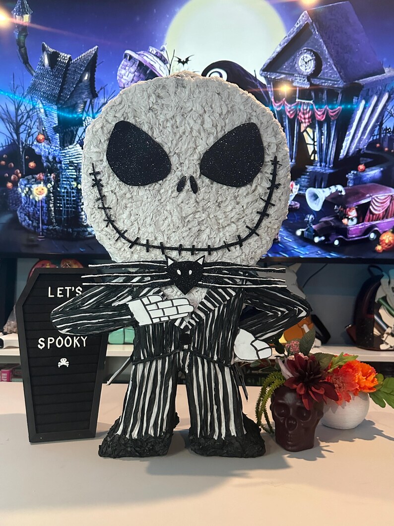 Puede incluir: Una pi&ntilde;ata de Jack Skellington en blanco y negro con un traje a rayas y una pajarita negra. La pi&ntilde;ata est&aacute; sobre una mesa con un letrero negro de pizarra que dice "Let's Spooky".