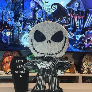 Puede incluir: Una pi&ntilde;ata de Jack Skellington a rayas blancas y negras con una pajarita negra y un gato negro en el pecho. La pi&ntilde;ata est&aacute; de pie sobre una mesa frente a una pizarra negra con las palabras "Let's Get Spooky" escritas en ella.