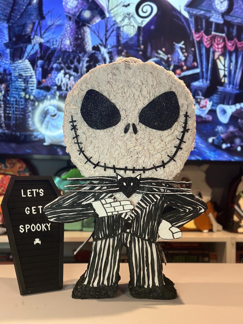 Puede incluir: Una figura de Jack Skellington con rayas blancas y negras, con una pajarita negra y un gato negro en el pecho. La figura est&aacute; de pie frente a una pizarra negra con el texto "Let's Get Spooky" escrito en ella.