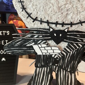 Puede incluir: Una pi&ntilde;ata a rayas blancas y negras de Jack Skellington de Pesadilla Antes de Navidad. La pi&ntilde;ata est&aacute; hecha de papel de seda y tiene una pajarita negra y una cara de gato negro en la parte delantera. La pi&ntilde;ata est&aacute; de pie sobre una superficie blanca.