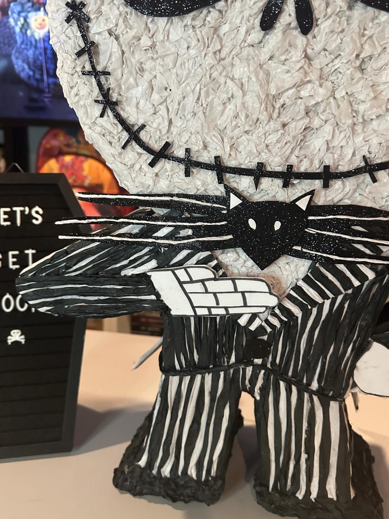 Puede incluir: Una pi&ntilde;ata a rayas blancas y negras de Jack Skellington de Pesadilla Antes de Navidad. La pi&ntilde;ata est&aacute; hecha de papel de seda y tiene una pajarita negra y una cara de gato negro en la parte delantera. La pi&ntilde;ata est&aacute; de pie sobre una superficie blanca.