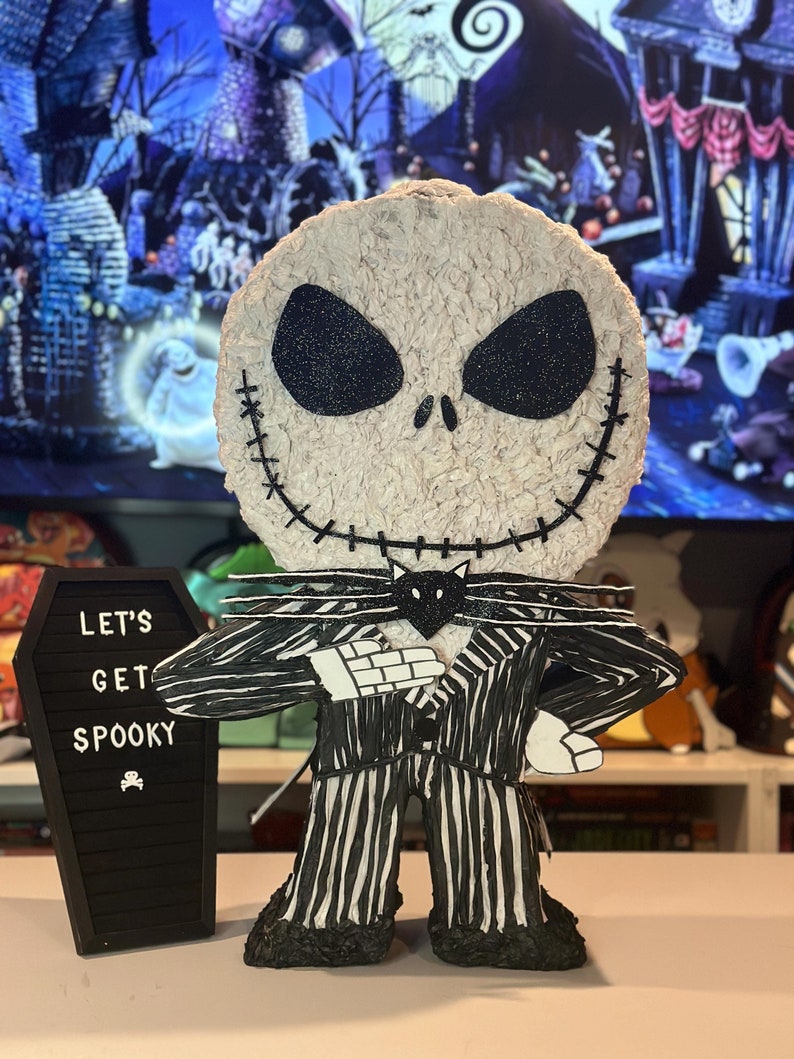 Puede incluir: Una pi&ntilde;ata de Jack Skellington a rayas blancas y negras con una pajarita negra y una cara de gato negro. La pi&ntilde;ata est&aacute; de pie sobre una mesa frente a un ata&uacute;d negro con las palabras "Let's Get Spooky" escritas en &eacute;l.