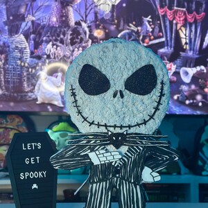 Puede incluir: Una figura de Jack Skellington con rayas blancas y negras y una pajarita negra. La figura sostiene un ata&uacute;d negro con el texto "Let's Get Spooky" escrito en blanco.