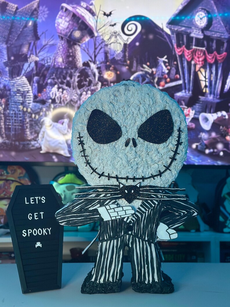 Puede incluir: Una figura de Jack Skellington con rayas blancas y negras y una pajarita negra. La figura sostiene un ata&uacute;d negro con el texto "Let's Get Spooky" escrito en blanco.
