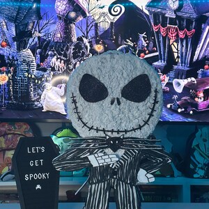 Puede incluir: Una figura de Jack Skellington con rayas blancas y negras y una pajarita negra. La figura est&aacute; de pie sobre una mesa con un letrero negro en forma de ata&uacute;d que dice "Let's Get Spooky".
