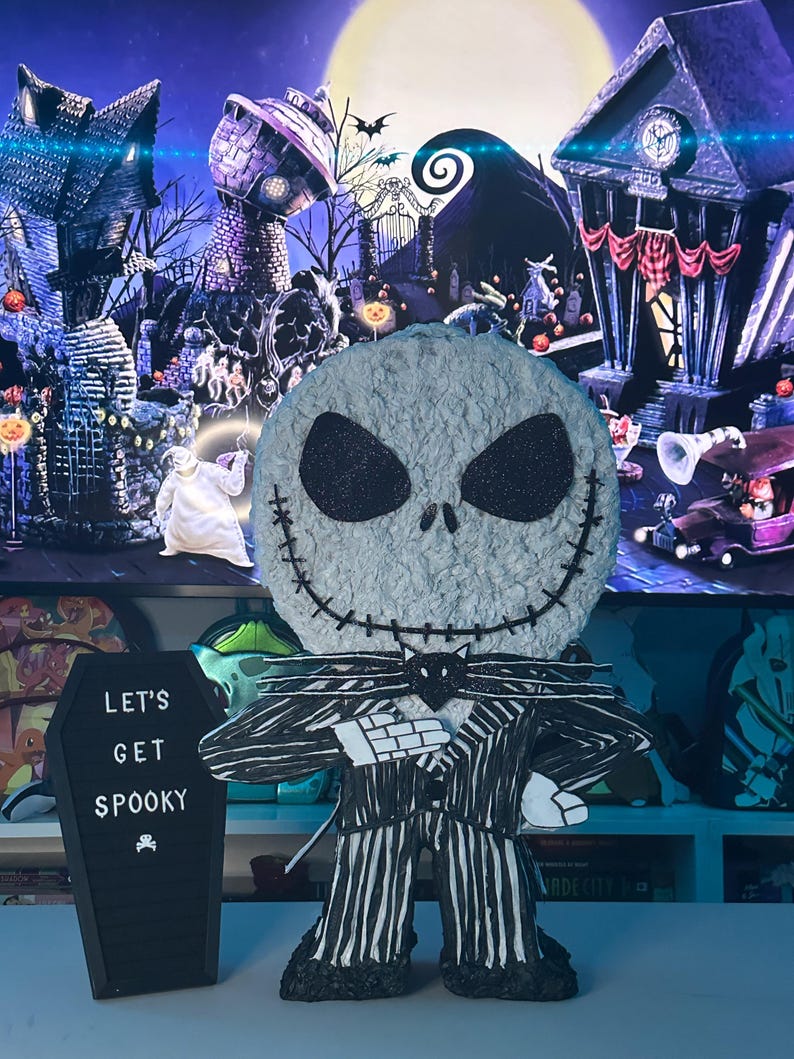 Puede incluir: Una figura de Jack Skellington con rayas blancas y negras y una pajarita negra. La figura est&aacute; de pie sobre una mesa con un letrero negro en forma de ata&uacute;d que dice "Let's Get Spooky".