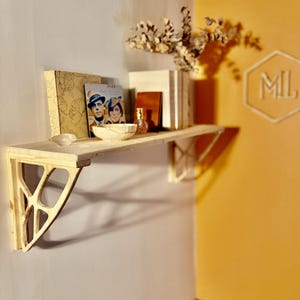 Pode incluir: Uma prateleira de madeira clara montada na parede, suportada por suportes decorativos. A prateleira contém livros, uma tigela pequena e outros itens decorativos. A parede é pintada em uma cor neutra, com uma seção de parede amarela visível.