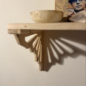 May include: A wooden shelf with a decorative bracket. The shelf is made of light-colored wood and features a carved bracket with a fan-like design. A light-colored wooden bowl sits on the shelf. The shelf is mounted on a white wall.