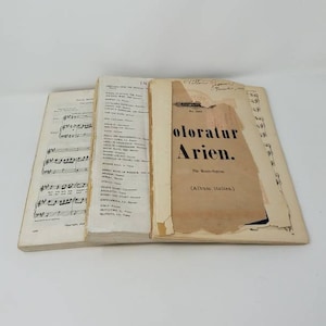 May include: Three vintage sheet music books with a white background. The book on the right has the title "Coloratur Arien." The book on the left has music notes and the text "Copyright MCMXXIX."