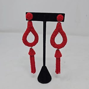 May include: A pair of red beaded earrings with a teardrop design and tassel detail. The earrings are displayed on a black stand against a white background. The earrings have a post backing.