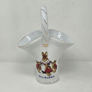 May include: White ceramic basket with a handle, featuring a gold trim and a colorful illustration of rabbits. The base of the basket reads "Mrs. Rabbit". A decorative piece for display or small items.