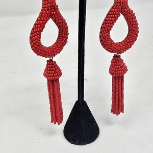 May include: Red beaded earrings featuring a teardrop shape and a tassel. The earrings are crafted from small, round beads and have a post fastening. They are displayed on a black stand against a white backdrop.
