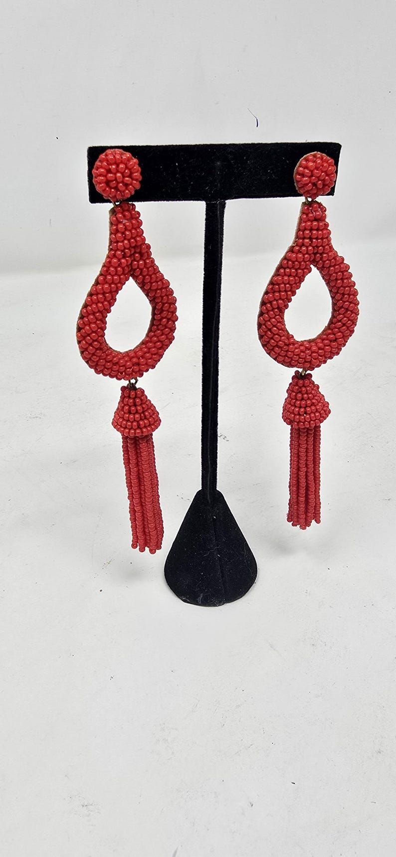 May include: Red beaded earrings featuring a teardrop shape and a tassel. The earrings are crafted from small, round beads and have a post fastening. They are displayed on a black stand against a white backdrop.