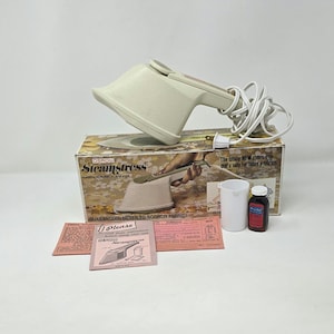 May include: An off-white Steamstress fabric steamer with a white cord, sitting on a box with the product name. The box includes the text "GUARANTEED NEVER TO SCORCH FABRIC!". Also included are a white cup, a small bottle, and instruction sheets.
