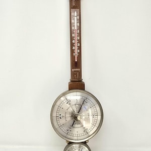 May include: Antique wooden barometer and thermometer combination. The thermometer displays temperature readings in Fahrenheit. The barometer has a silver face with a needle and numbers indicating atmospheric pressure. A decorative silver eagle tops the piece.