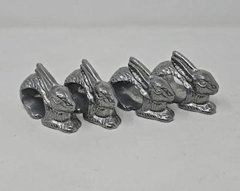 May include: Set of four silver-colored metal napkin rings. Each ring features a detailed bunny rabbit design. The rabbits are in a sitting position with their ears perked up. These rings are ideal for Easter or spring table settings.