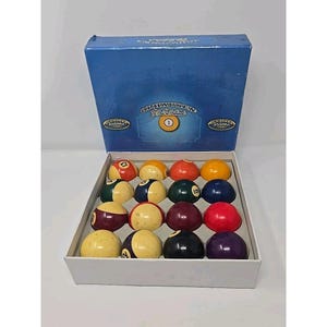 Può includere: Un set completo di palle da biliardo in una scatola bianca, con coperchio blu. Le palle sono di vari colori, tra cui giallo, rosso, verde e nero, ognuna con un numero. La scatola ha il testo "Olhausen Billiards".