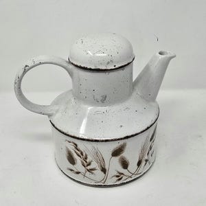 May include: White enamel teapot with a rounded body, spout, and handle. The teapot features a lid and is decorated with brown wheat stalks. The edges of the teapot are outlined in brown, giving it a vintage look.