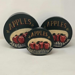 Op de afbeelding: Drie ronde, houten decoratieve stukken met een donkergroene achtergrond. Elk stuk heeft het woord "APPLES" in rood schrift boven een geschilderde scène van rode appels. De appels zijn gerangschikt op een gestreept oppervlak.