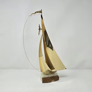 May include: A brass sailboat sculpture with a flag and a bird, mounted on a marble and wood base. The sailboat's sails are curved, and a thin wire arcs from the top of the mast. The sculpture is approximately 12 inches tall.