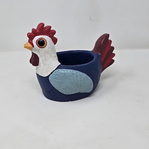 May include: A ceramic chicken-shaped planter. The planter is primarily navy blue with a white head, red comb and tail feathers, and a light blue wing. The beak is yellow, and the eyes are black.