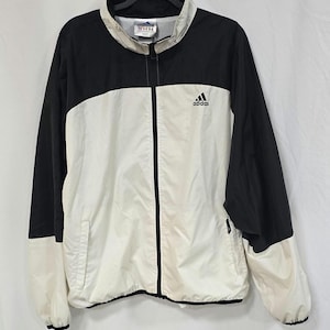 以下が含まれることがあります： 黒と白のアディダス製トラックジャケット。フルジッパー付き。襟と袖は黒、ボディは白。胸にアディダスのロゴ。黒いハンガーにかかっています。