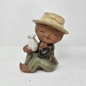 Może przedstawiać: Ceramiczna figurka siedzącej postaci w słomkowym kapeluszu, trzymającej mały biały dzbanek. Postać ubrana jest w zielone spodnie i brązową kurtkę. Figurka ma uśmiechniętą minę i prawdopodobnie jest przedmiotem dekoracyjnym.