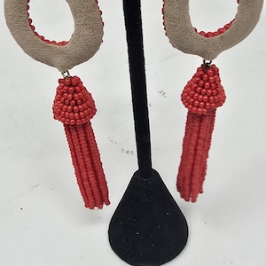 May include: A pair of teardrop earrings. They have a light brown suede-like material with a red beaded border. A cluster of red beads hangs from the bottom of each earring, forming a tassel.