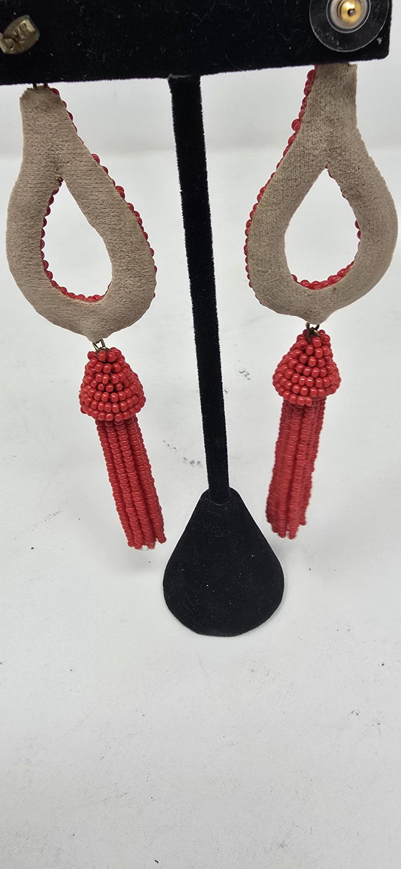 May include: A pair of teardrop earrings. They have a light brown suede-like material with a red beaded border. A cluster of red beads hangs from the bottom of each earring, forming a tassel.