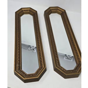 May include: A pair of tall, narrow mirrors with elongated octagonal frames. The frames are finished in a muted gold tone and feature a decorative, repeating embossed pattern along the inner edge surrounding the reflective glass surface.