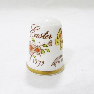 Puede incluir: Dedal de porcelana blanca con ribete dorado. Tiene un diseño floral con una mariposa y las palabras "Easter 1979" impresas en él.