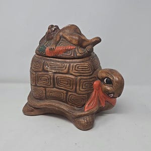 Könnte beinhalten: Braune Keramik-Schatulle in Schildkrötenform mit Deckel, der einen Hasen mit einer Karotte zeigt. Die Schildkröte hat ein strukturiertes Schilddesign und einen Hasenkopf mit roter Schleife. Die Box ist ein dekorativer Artikel.