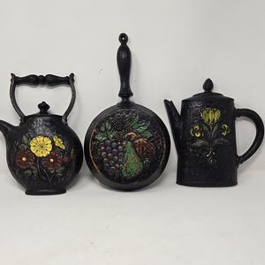 May include: Three black decorative kitchen items: a kettle, a frying pan, and a teapot. The kettle and teapot feature floral designs in yellow and red. The frying pan has a fruit design with grapes and pears.
