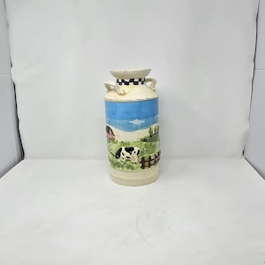 May include: A ceramic milk can-shaped vase with a farm scene. The vase features a cream-colored top and base, a blue sky, and a green pasture with a cow and a fence. The vase has two handles.