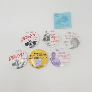 May include: A collection of nine colourful round buttons featuring various events and performers. The buttons are all white with black text and images. The events include "Dvoika!" "Footprints on the Moon", "Obo Addy & Kukrudu", "Ruth Golden", and "Jackie Torrence".