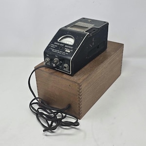 May include: A black Grid-Dip Meter, model 90651, with a white dial and various control knobs, sits atop a wooden box. The meter has a label that reads "James Millen Mfg. Co., Inc. Malden, Mass. U.S.A." A black cord is connected to the meter.
