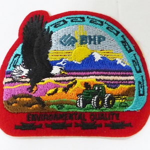 May include: A red embroidered patch with a blue and green border. The patch features a bald eagle, a sun, mountains, and a tractor. The text "Environmental Quality" is embroidered at the bottom of the patch.