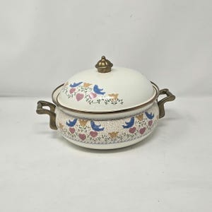 May include: Cream-colored enamelware casserole dish with lid. The dish features a decorative pattern of blue birds, pink hearts, and yellow flowers. It has brass-colored handles and a knob on the lid.