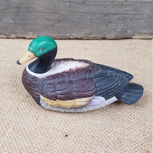 Puede incluir: Una figurita de pato de cerámica con la cabeza verde, plumas marrones y negras y la parte inferior blanca. El pato está acostado boca abajo con la cabeza girada hacia un lado.