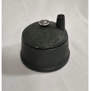 May include: A black cast iron pencil sharpener with a textured, matte finish. The cylindrical body features a removable lid with a small, centered silver-toned metal knob and a single vertical opening for inserting pencils at the top edge.