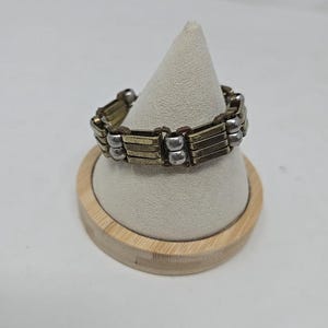 May include: A stretch bracelet with alternating gold and silver-toned rectangular links. The bracelet is displayed on a white cone-shaped stand with a wooden base. The jewelry is designed to be worn on the wrist.