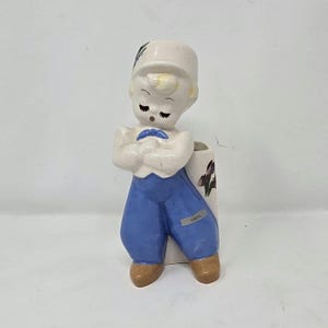 May include: A ceramic figurine of a boy in Dutch attire, featuring a white hat, blue pants, and a white top. The figurine has a vase-like opening in the back and is decorated with floral accents. The word "HAND" is on the pants.