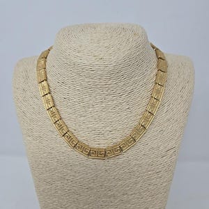 May include: A gold-toned necklace with a geometric Greek key design. The necklace is displayed on a beige mannequin bust. The necklace is composed of rectangular links, each featuring the repeating key pattern. The jewelry is a statement piece.